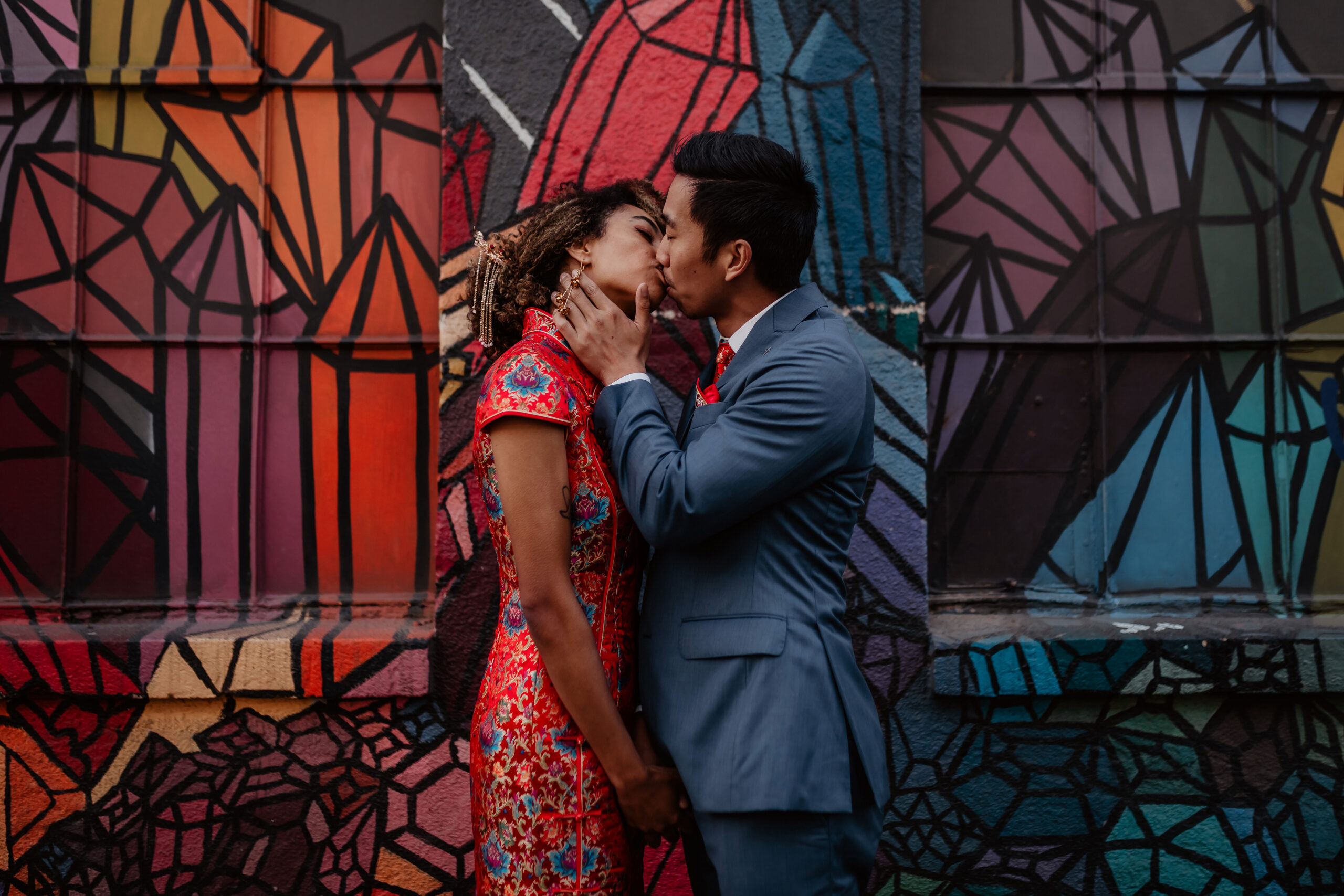 Couple in Chinese wedding clothes kissing in front of a colorful mural in Capitol Hill
